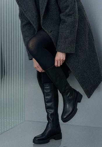 Zwarte kniehoge laarzen met een glad lederen textuur, vierkante neus en stevige blokhak, gecombineerd met zwarte panty's en een getextureerde grijze jas.
