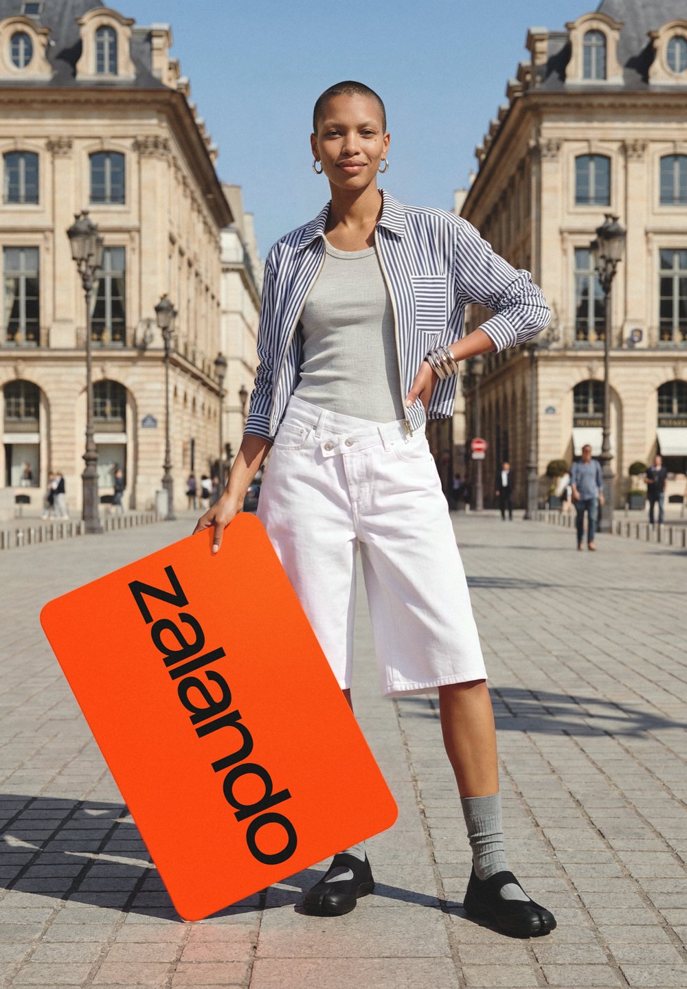 Person steht draußen auf dem Stadtplatz und hält eine große, leuchtend orange Zalando-Karte, trägt weiße Shorts, ein gestreiftes Hemd, ein graues Top und schwarze Schuhe.