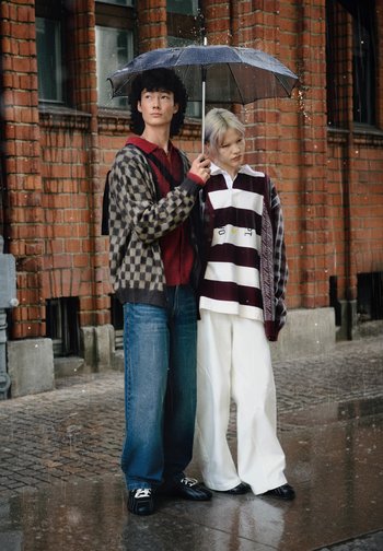 Two individuals stand under a black umbrella in the rain. One wears a checkered cardigan, red sweater, and blue jeans; the other, a striped shirt and white pants.