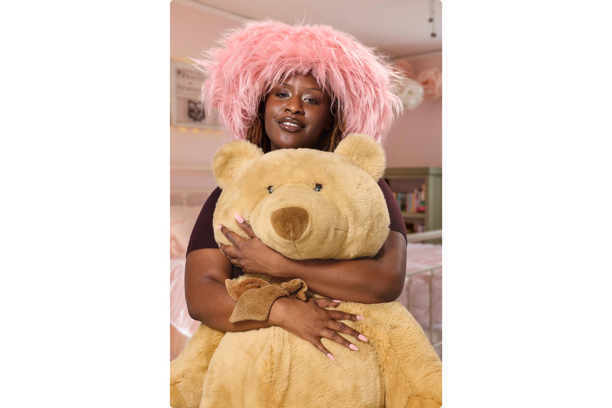 Una donna con i capelli lunghi e un cappello rosa morbido abbraccia un grande orsetto di peluche in una stanza decorata con delicatezza.
