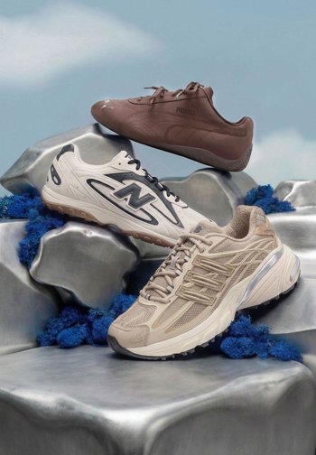 Tres zapatillas deportivas flotantes: una Puma marrón, una New Balance blanca y una New Balance beige, mostradas sobre rocas plateadas con musgo azul.