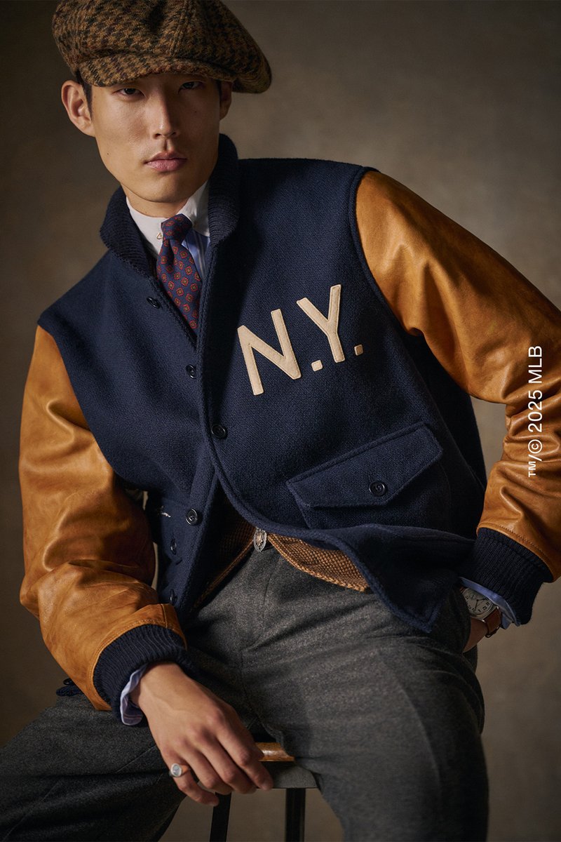 Giacca di lana navy con maniche in pelle tan e lettering "N.Y.". Presenta chiusure a bottone e un interno testurizzato, abbinata a pantaloni grigi.