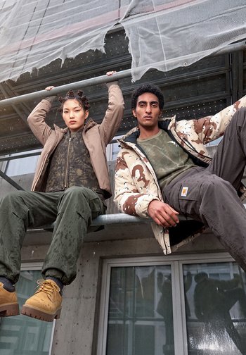 Deux mannequins sont assis sur un échafaudage métallique devant un bâtiment. Ils portent des vestes d’extérieur, des pantalons cargo et des chaussures de travail, sous un filet de sécurité déchiré.