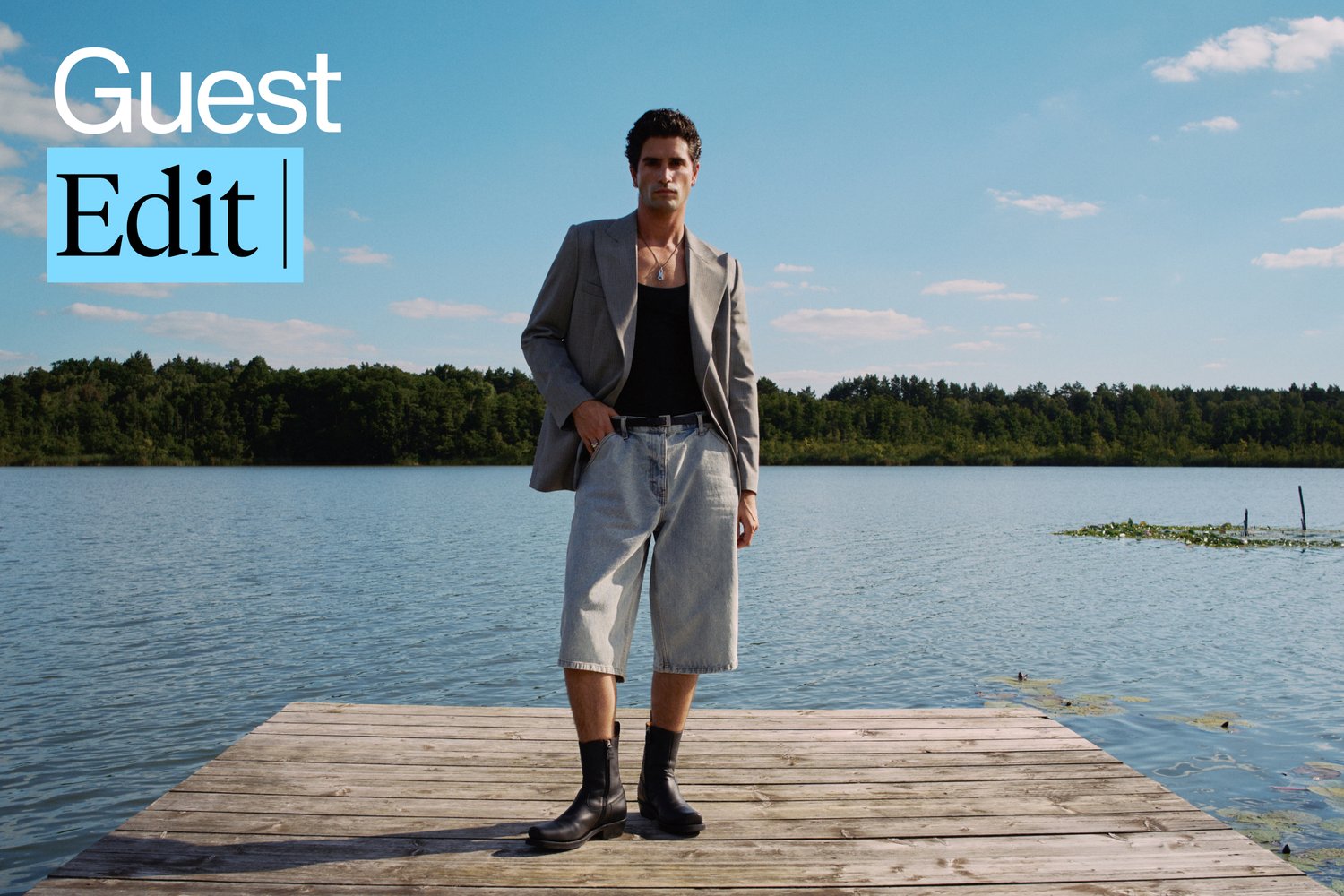 A person stands on a wooden pier by a lake, wearing a blazer, tank top, and denim shorts, with green trees and blue sky in the background.