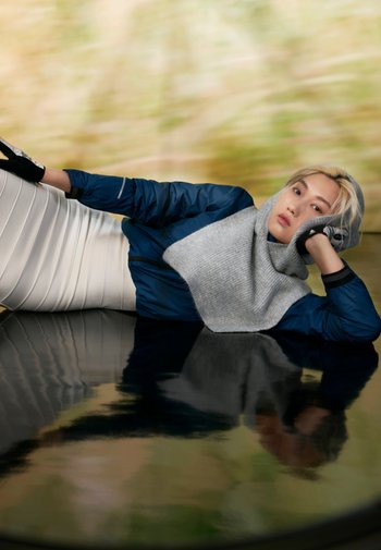 Eine Person mit hellen Haaren, die einen grauen Schal und eine dunkle Jacke trägt, lehnt sich auf einer reflektierenden Fläche zurück, mit einem abstrakten, verschwommenen Hintergrund.