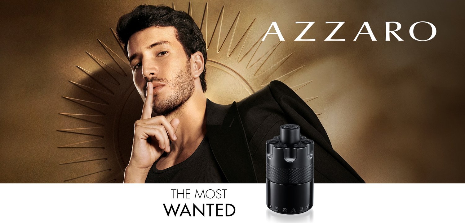 Homme avec barbe taillée et veste noire faisant un geste de silence à côté d'un flacon de parfum Azzaro noir sur un fond doré.
