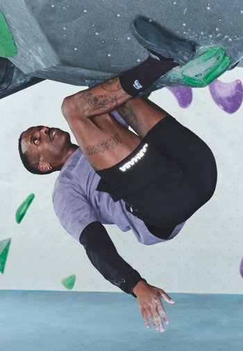 Alpinist îmbrăcat într-un tricou mov cu mânecă scurtă și pantaloni scurți negri, ținându-se de prizele unui perete de escaladă cu elemente verzi și mov, cu mâinile pudrate cu magneziu.