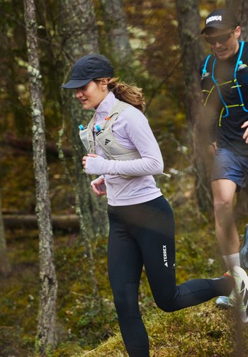 To løpere med hatter og væskeryggsekker løper på sti gjennom en skog med mose dekket bakken og trær.