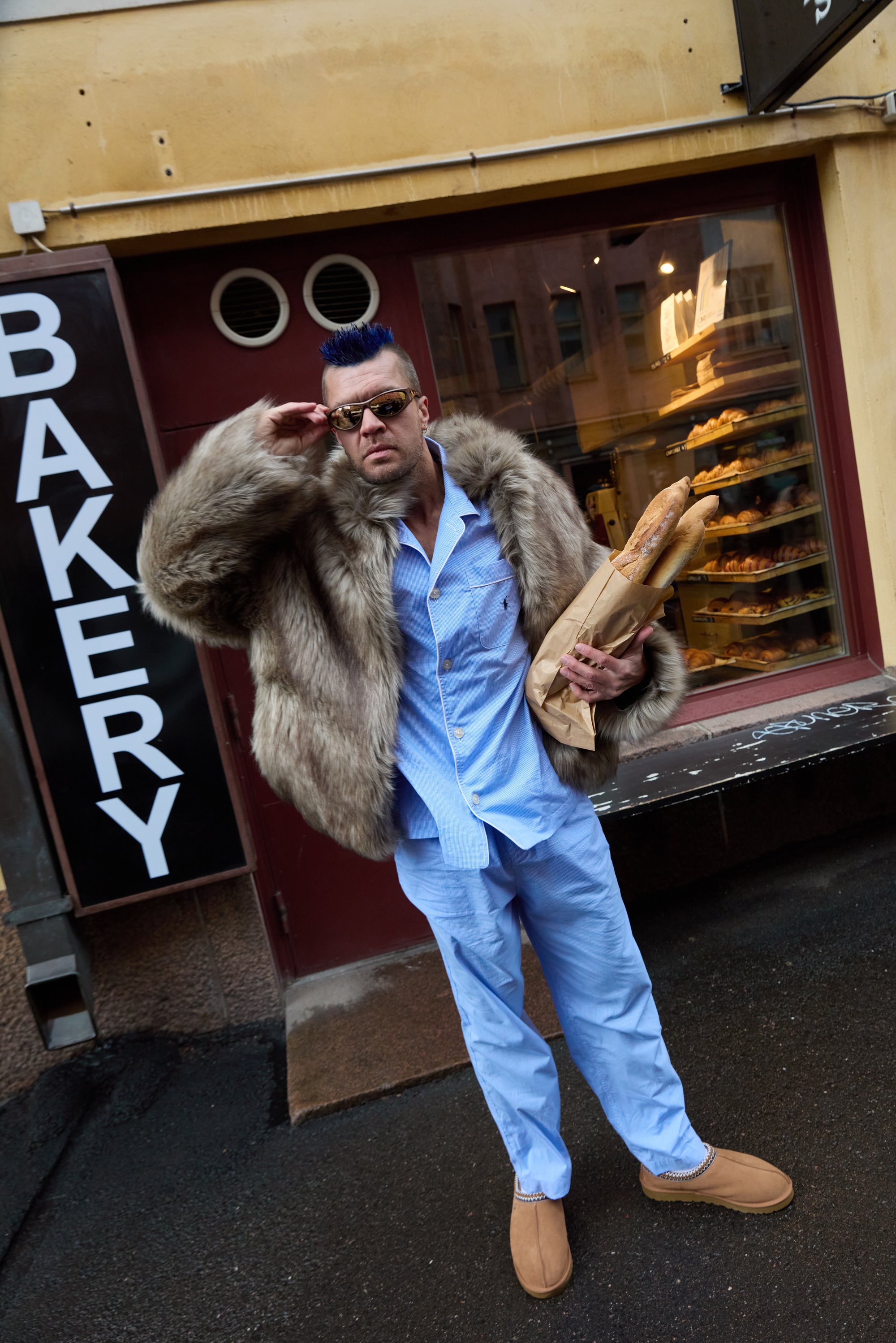 Tekoturkistakki vaaleanruskeana, vaaleansininen pyjama-asu, vaaleanruskeat tohvelit, aurinkolasit, kantaa leipomon ulkopuolella pusseihin pakattuja patonkeja kyltein varustetun leipomon edessä.