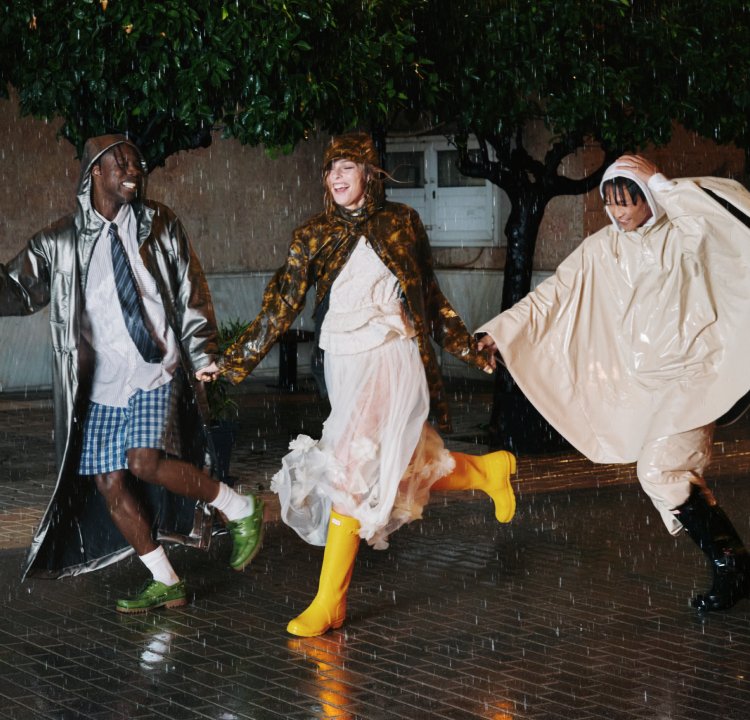 Trois personnes en imperméables et bottes dansant joyeusement dans une rue mouillée la nuit sous la pluie, se tenant la main et souriant.