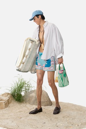 A person wearing a baseball cap and sunglasses carries a folding chair and striped bag, dressed in beachwear, on sandy terrain.
