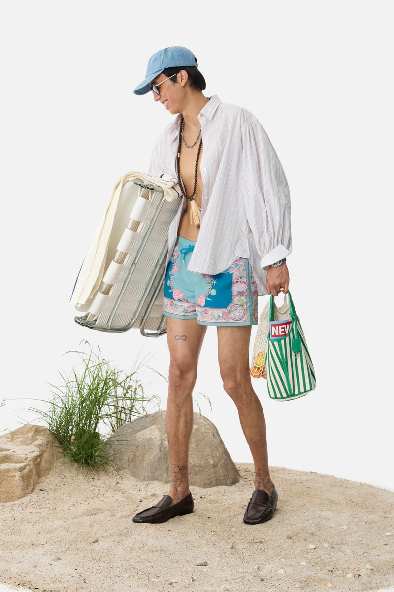 Una persona con un cappellino da baseball e occhiali da sole porta una sedia pieghevole e una borsa a righe, vestita con abbigliamento da spiaggia, su terreno sabbioso.