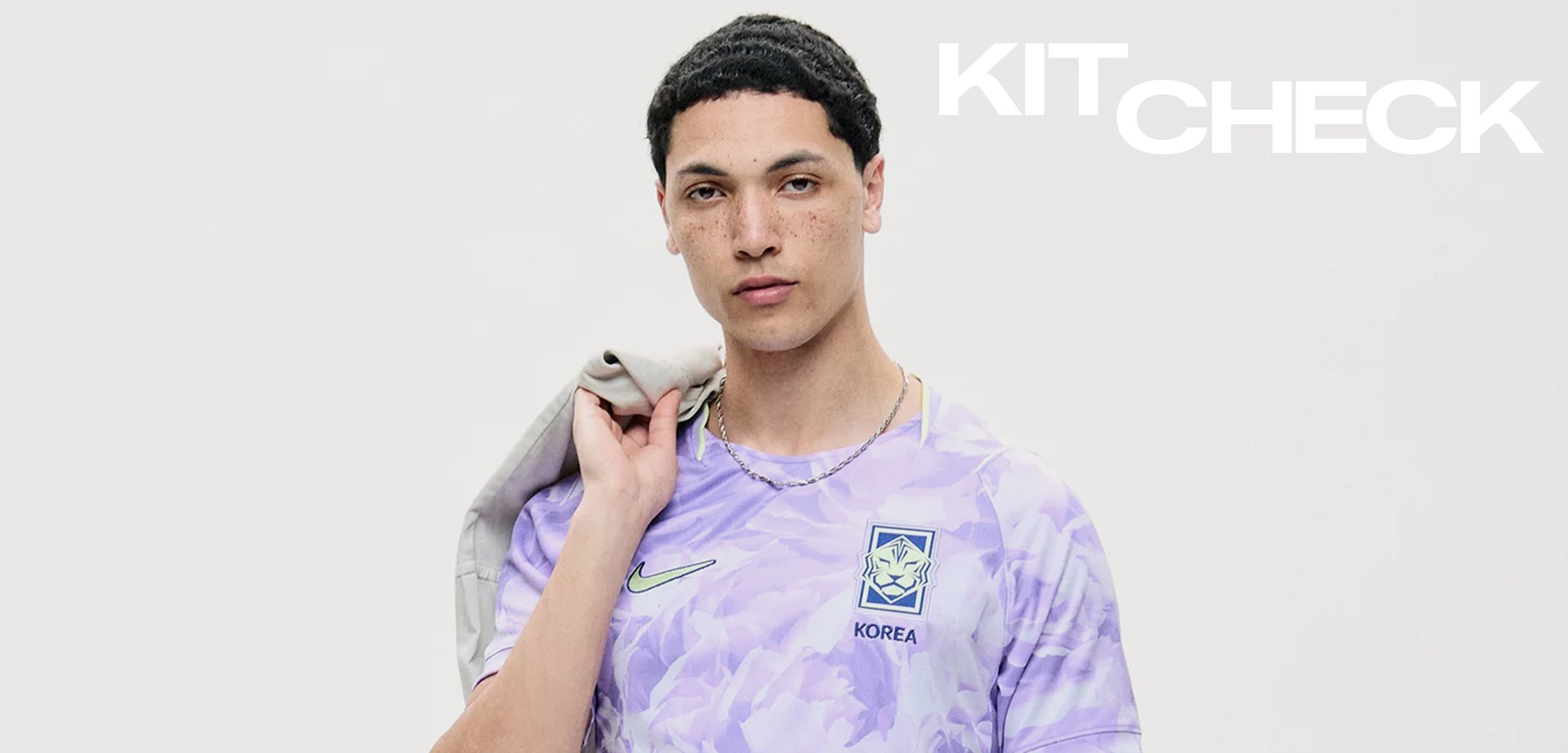 Junger Mann in einem violetten Südkorea Fussballtrikot, der eine graue Jacke über der Schulter trägt und vor einem schlichten hellen Hintergrund steht.