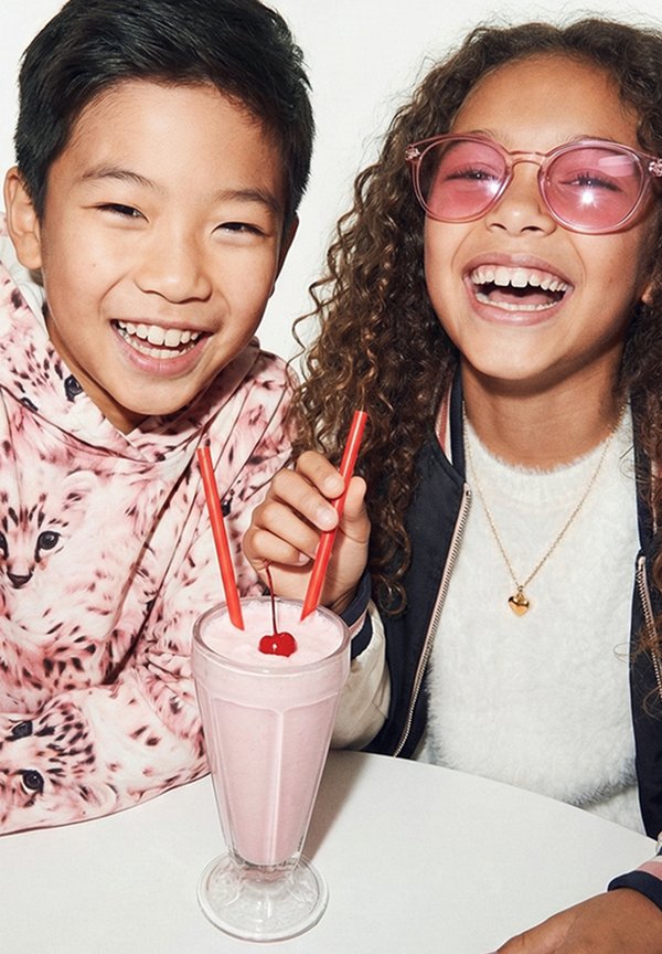 Dos niños sonriendo y compartiendo un batido rosa con pajillas rojas y una cereza encima en una mesa blanca.