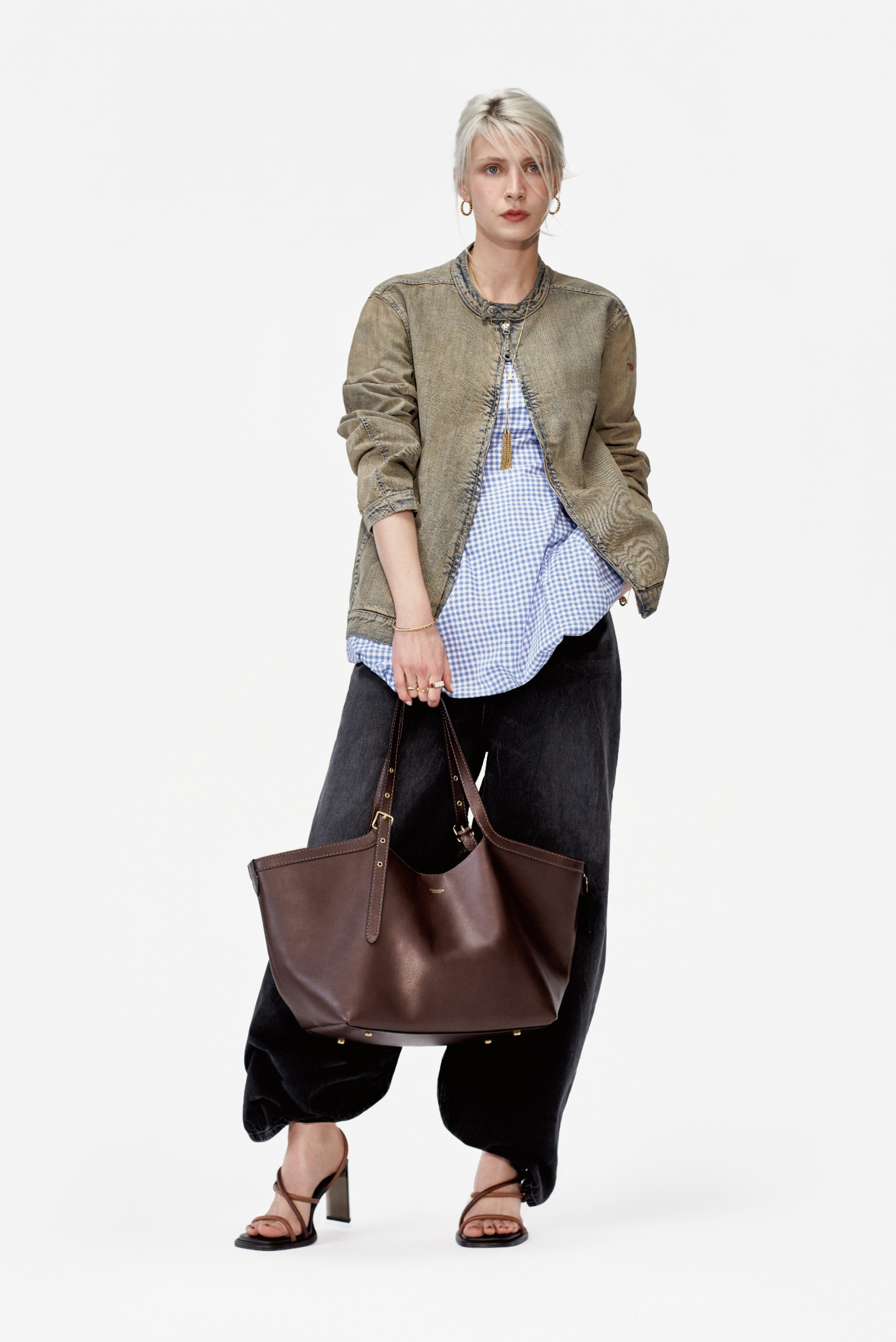 Una mujer con cabello corto, rubio platino, lleva una blusa de cuadros azul claro, una chaqueta bomber oliva y pantalones de pierna ancha, sosteniendo un bolso marrón.
