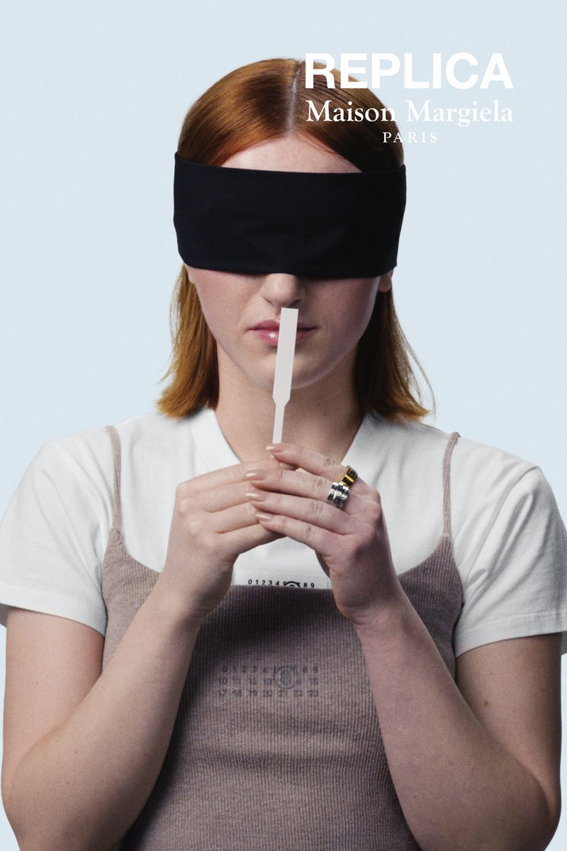 Black blindfolded figure holds a white fragrance strip. They wear a white shirt under a beige ribbed dress. Background is light blue.