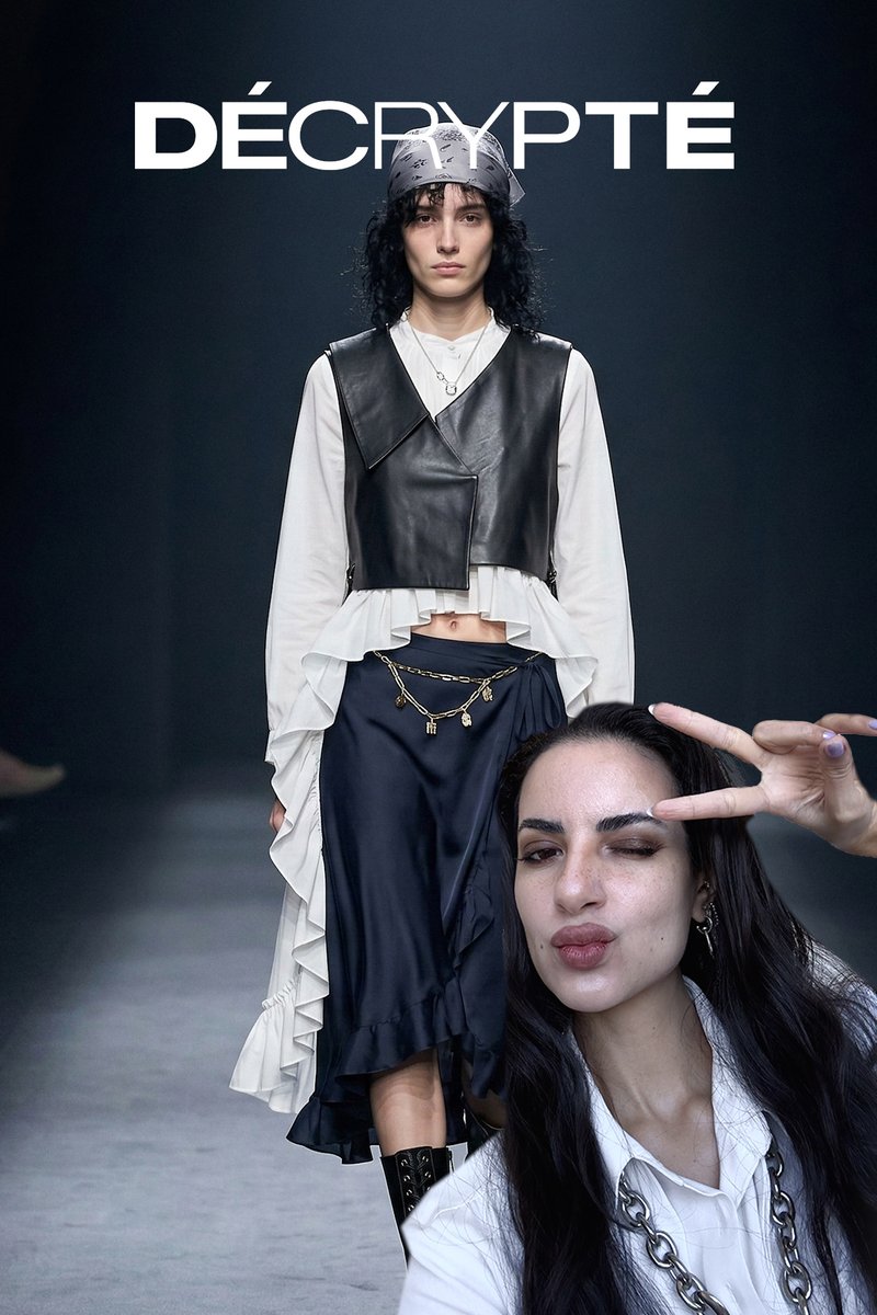 Un mannequin féminin défile sur le podium dans une tenue superposée noir et blanc avec un gilet en cuir et une ceinture chaîne, tandis qu'une autre femme fait un signe de paix et un clin d'œil au premier plan.