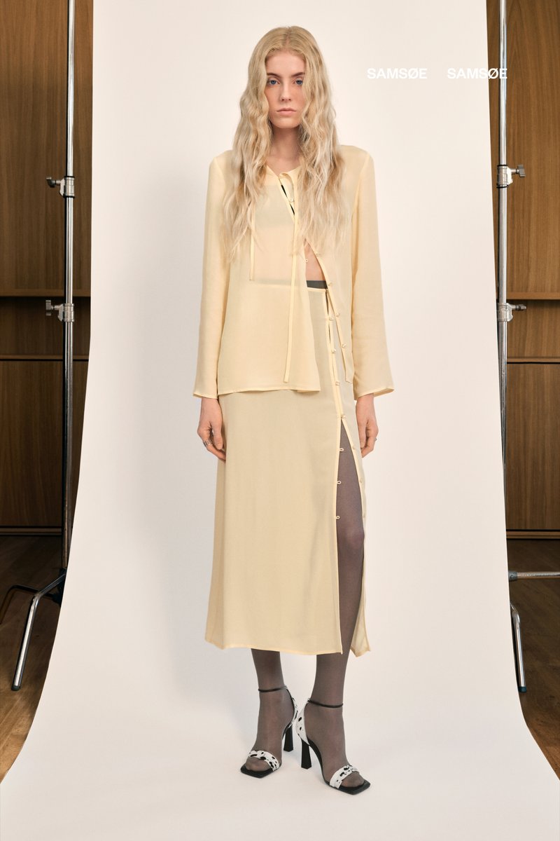 Helles gelbes, transparentes Oberteil mit langen Ärmeln und einer Bindeschließe, kombiniert mit einem passenden Rock mit seitlichem Schlitz und Knöpfen. Schwarz-weiße High Heels.