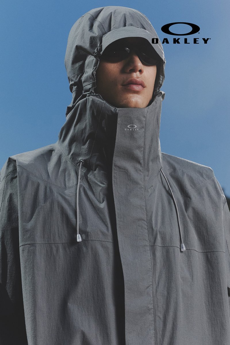 Una persona indossa una giacca grigia Oakley con cappuccio e occhiali da sole, sullo sfondo di un cielo blu sereno. Il logo del marchio è visibile nella parte superiore.