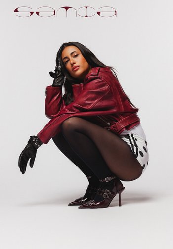 Femme accroupie dans une veste en cuir bourgogne, des gants noirs, des collants noirs transparents, un short à pois et des talons assortis bourgogne, sur fond blanc uni.