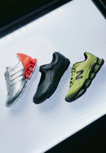 Drei verschiedene Sportschuhe nebeneinander auf einer beleuchteten weißen Oberfläche: silber und orange, schwarz und grün mit schwarzen Sohlen.