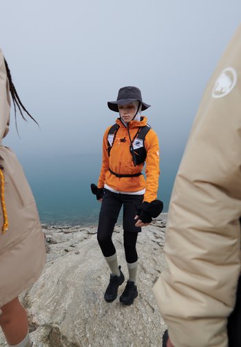 Eine Person in einer orangefarbenen Jacke und einem Hut bewegt sich über felsiges Terrain in der Nähe eines ruhigen Gewässers, umgeben von nebligen Bedingungen.
