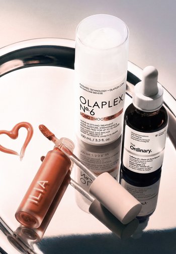 Gloss à lèvres avec applicateur, flacon de traitement capillaire Olaplex No.6 et sérum visage The Ordinary sur un plateau argenté réfléchissant.