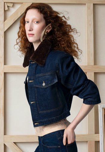Femme aux cheveux roux bouclés portant une veste en denim foncé avec col en fourrure, un haut beige en dentelle, et une grosse boucle d'oreille chaîne en or, debout de profil.