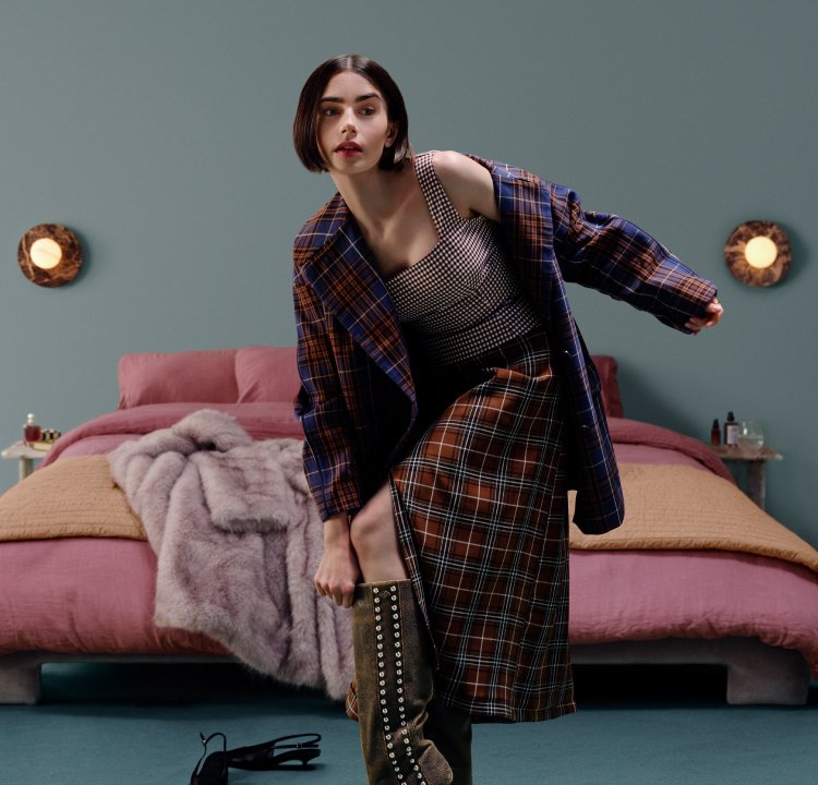 Vrouw in ruitjas en rok die haar hoge laarzen met studs afstelt terwijl ze in een slaapkamer staat met roze en beige beddengoed.