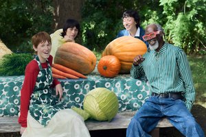 Cuatro personas están sentadas alrededor de una mesa con un mantel de estampado floral. Están rodeadas de grandes calabazas, zanahorias y un repollo.