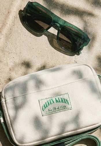 Green translucent sunglasses resting beside a beige Calvin Klein crossbody bag with green text and strap on a stone surface.