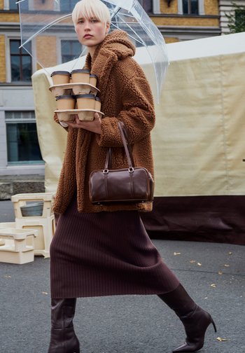 Brun, fluffy, overdimensioneret frakke; ribbet, mørkebrun nederdel; brun lædertaske; klar paraply; bærer en bakke med fem kaffekopper.