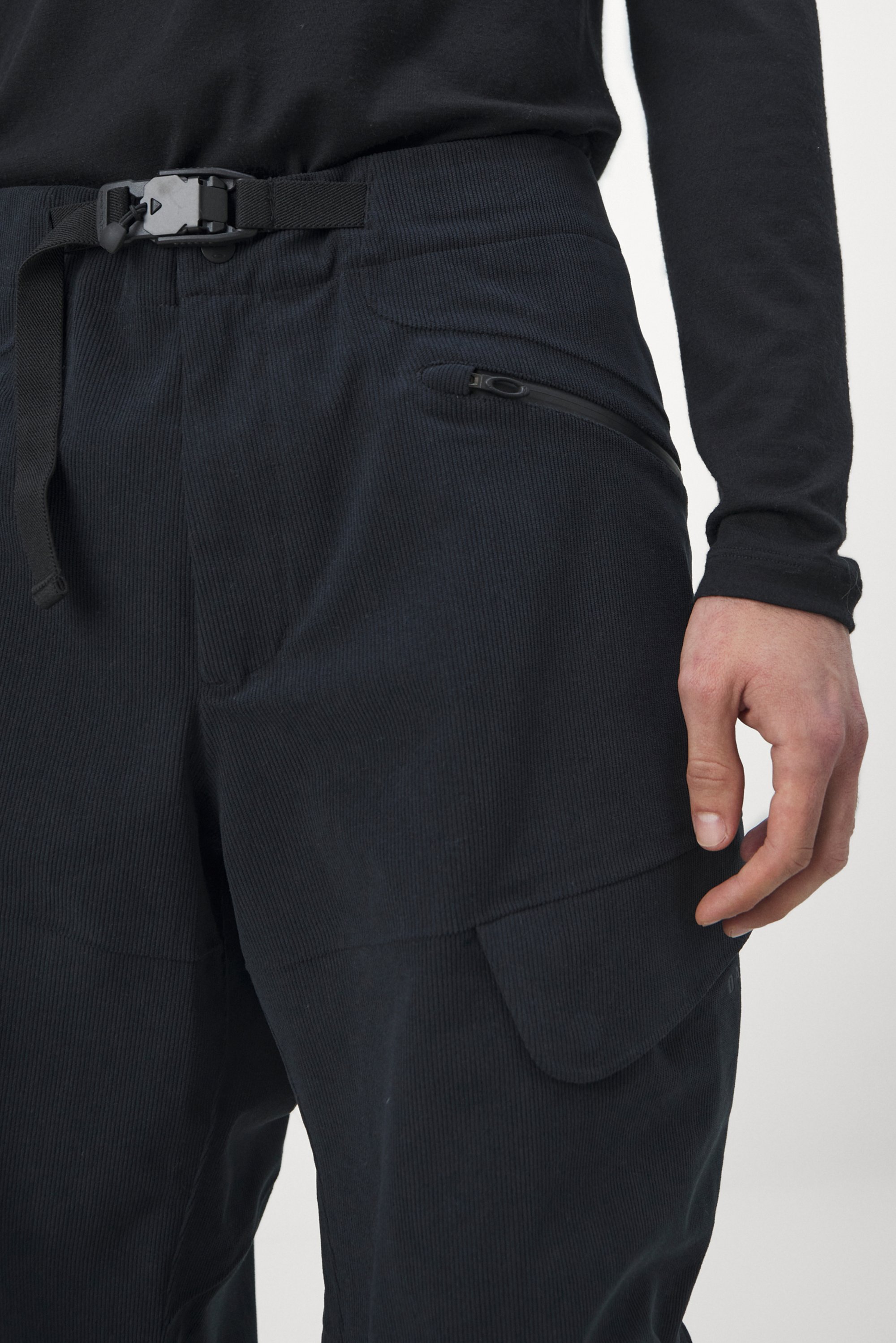 Dark, textured pants with a belt and zippered pocket, shown from the waist down, with a hand resting near the pocket.