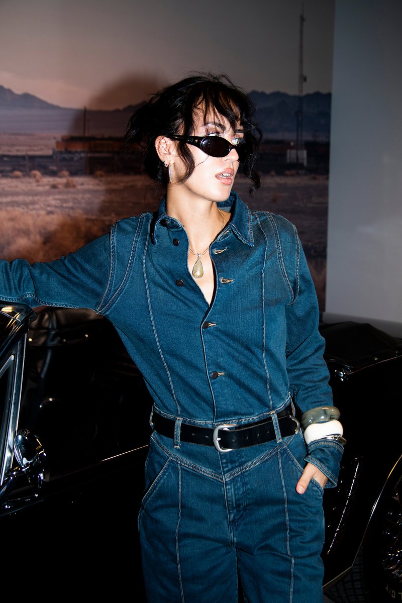 Denim jumpsuit in donkerblauw met knopen aan de voorkant, lange mouwen en een getailleerde taille. Accessoires zijn onder meer dikke armbanden en donkere zonnebrillen.