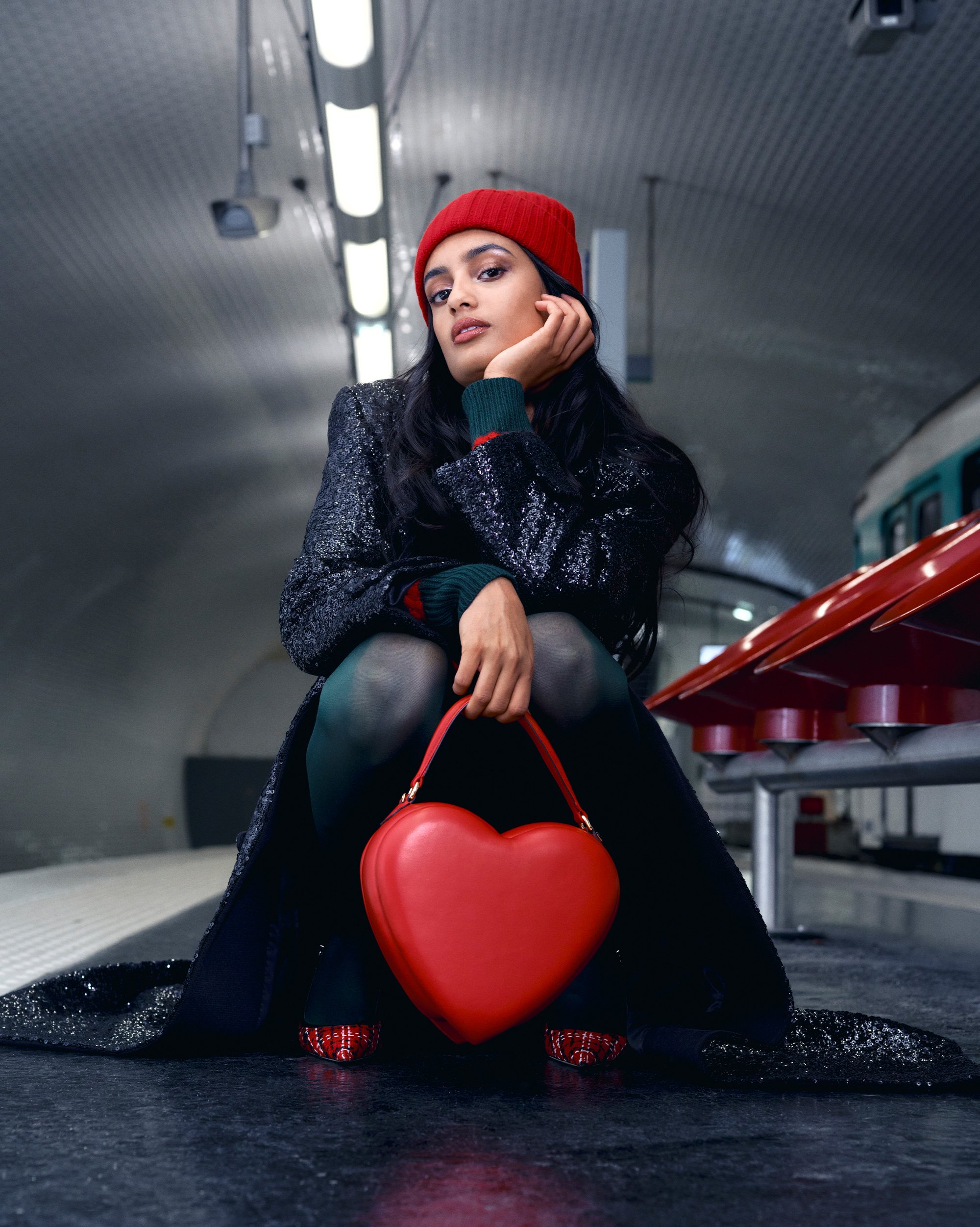 A woman with long dark hair sits on a subway platform, wearing a red beanie and black sequined coat, holding a heart-shaped red handbag.