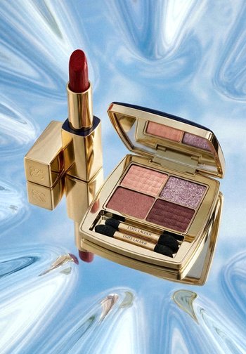 Red lipstick with gold case and open gold eyeshadow palette showing four shades and two applicators on blue satin fabric background.
