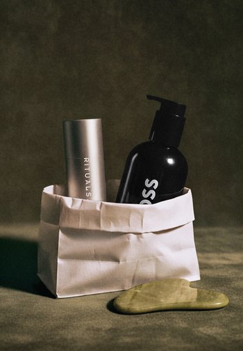 Deux flacons cosmétiques, un argent et un noir avec une pompe, à l'intérieur d'un sac en papier blanc plié, à côté d'une pierre verte plate sur une surface marron.