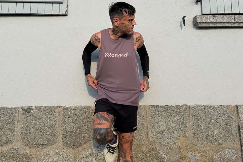Junger Mann mit Tattoos, der ein ärmelloses Shirt mit der Aufschrift "Normal" und schwarze Shorts trägt, steht vor einer Stein- und weißen Wand.