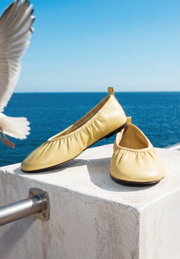 Chaussons de ballet en cuir jaune clair sur un rebord en béton blanc avec océan bleu et aile d’oiseau blanche volante en arrière-plan.