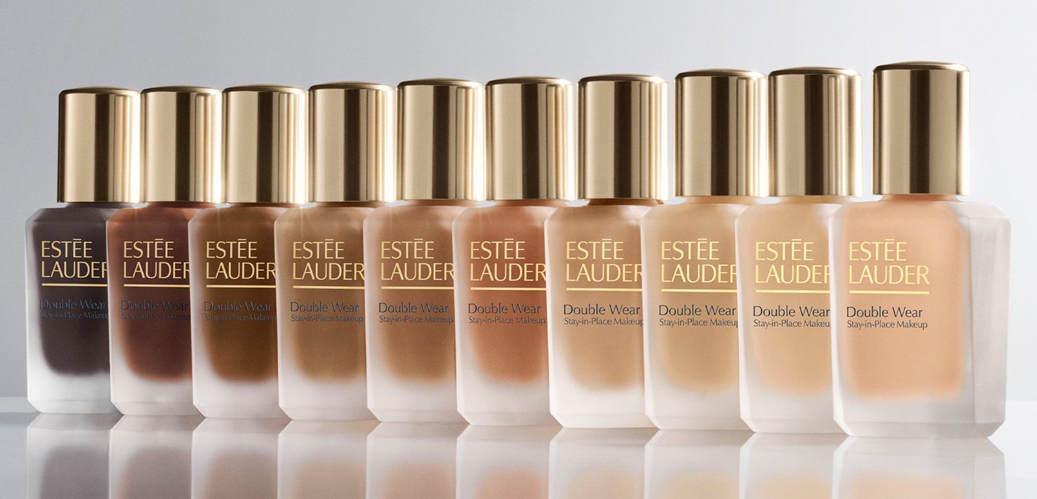 Doce botellas de base Estée Lauder Double Wear en fila, mostrando un degradado de tonos de piel desde oscuro hasta claro, con tapas doradas.