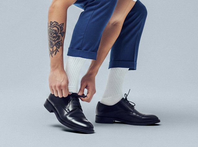 Svarta läderskor med rund tå och snörning, matchade med vita ribbade strumpor och blå byxor. Märkbart tatuering på vänster arm.