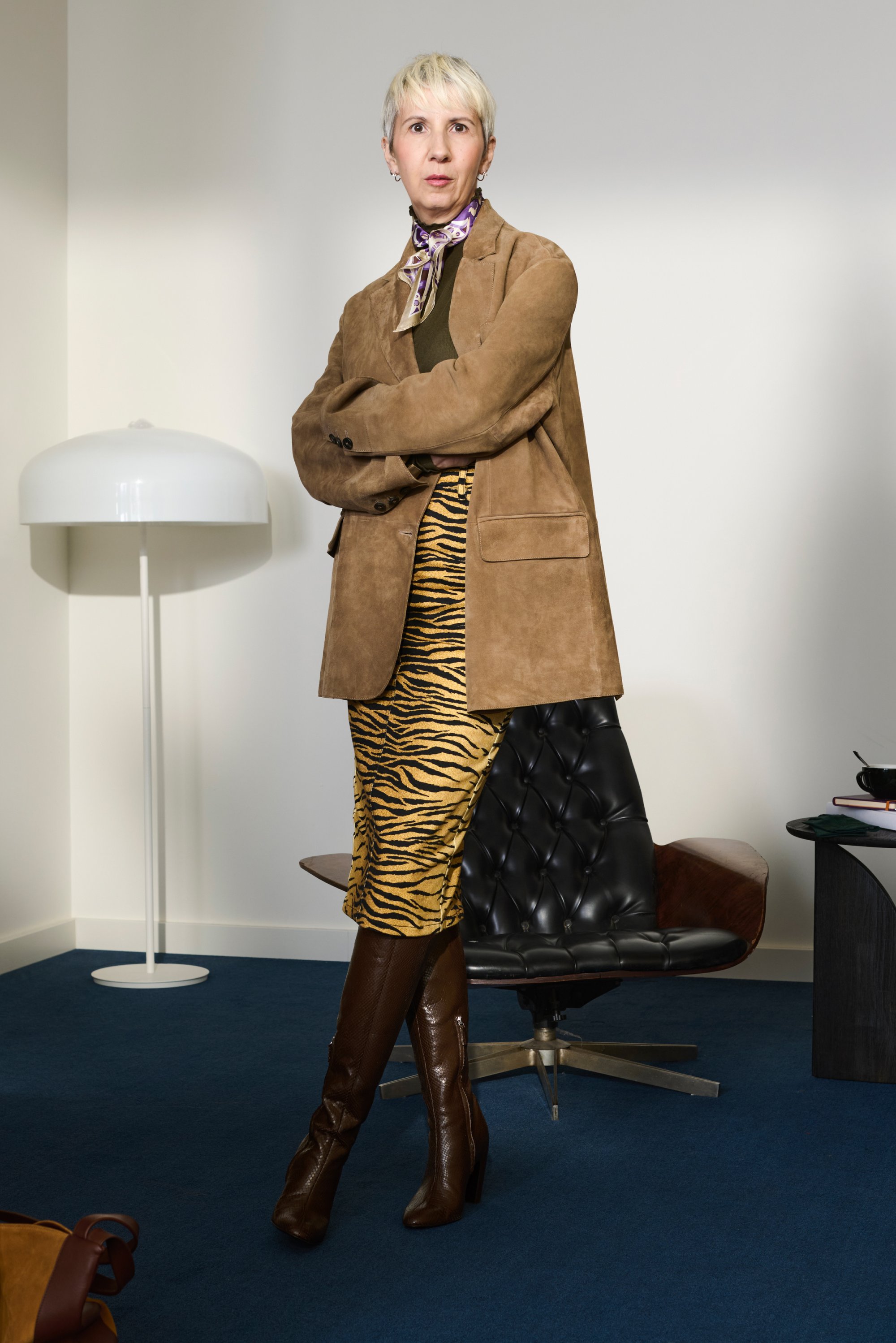 Blazer in suede color tan sopra un top oliva, abbinato a una gonna a strisce gialle e nere e stivali marroni fino al ginocchio. Sedia nera con puff e lampada nello sfondo.