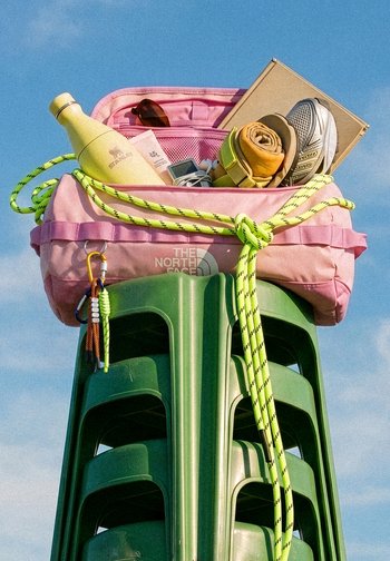 Pinkfarbene Sporttasche auf gestapelten grünen Plastikstühlen enthält Schuh, zusammengerolltes Handtuch, Wasserflasche, Sonnenbrille, Notizbuch und Ohrhörer vor blauem Himmel.