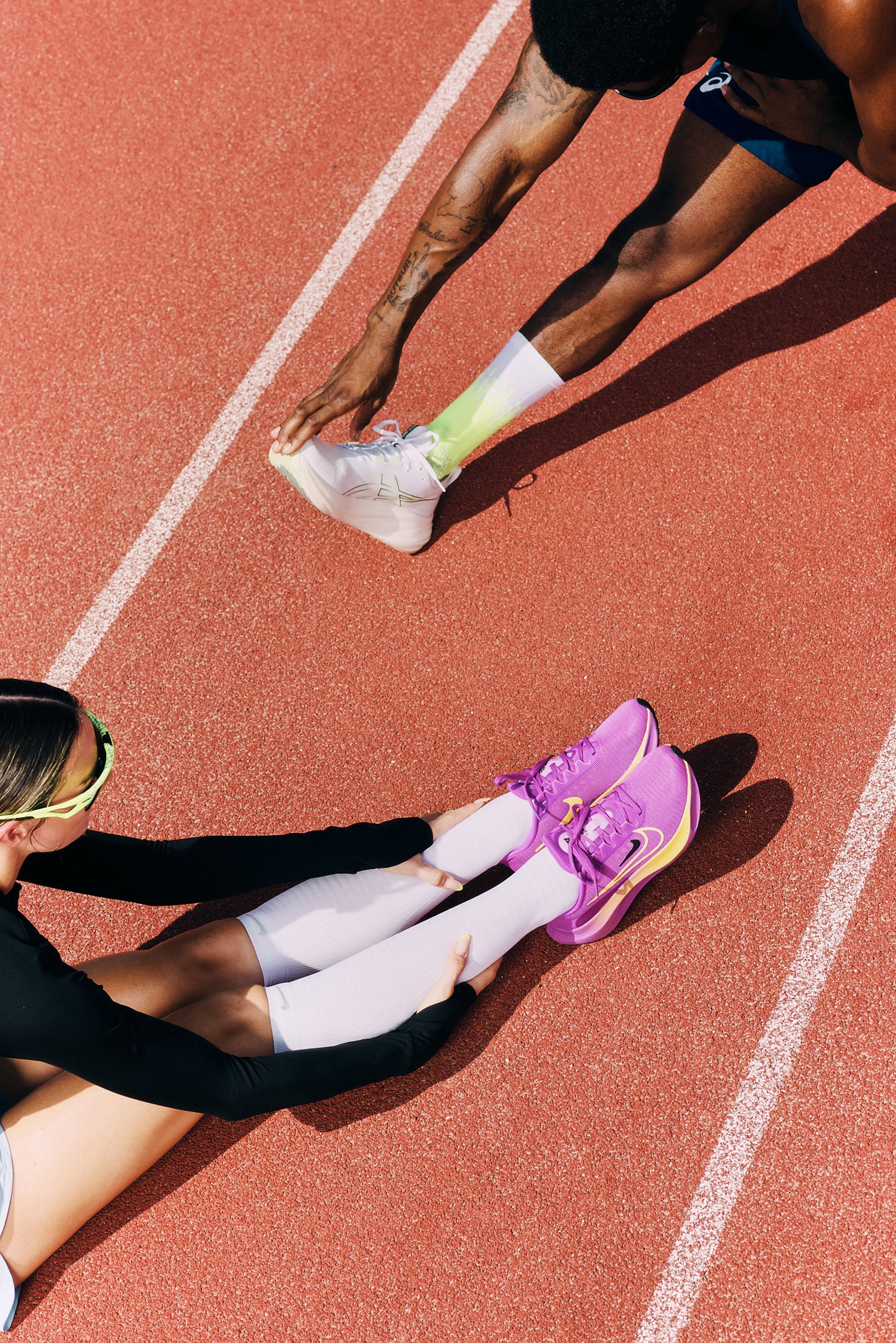 A woman stretches her legs on a track, wearing black long sleeves and white leggings. A man nearby stretches his leg, wearing green socks and shoes.