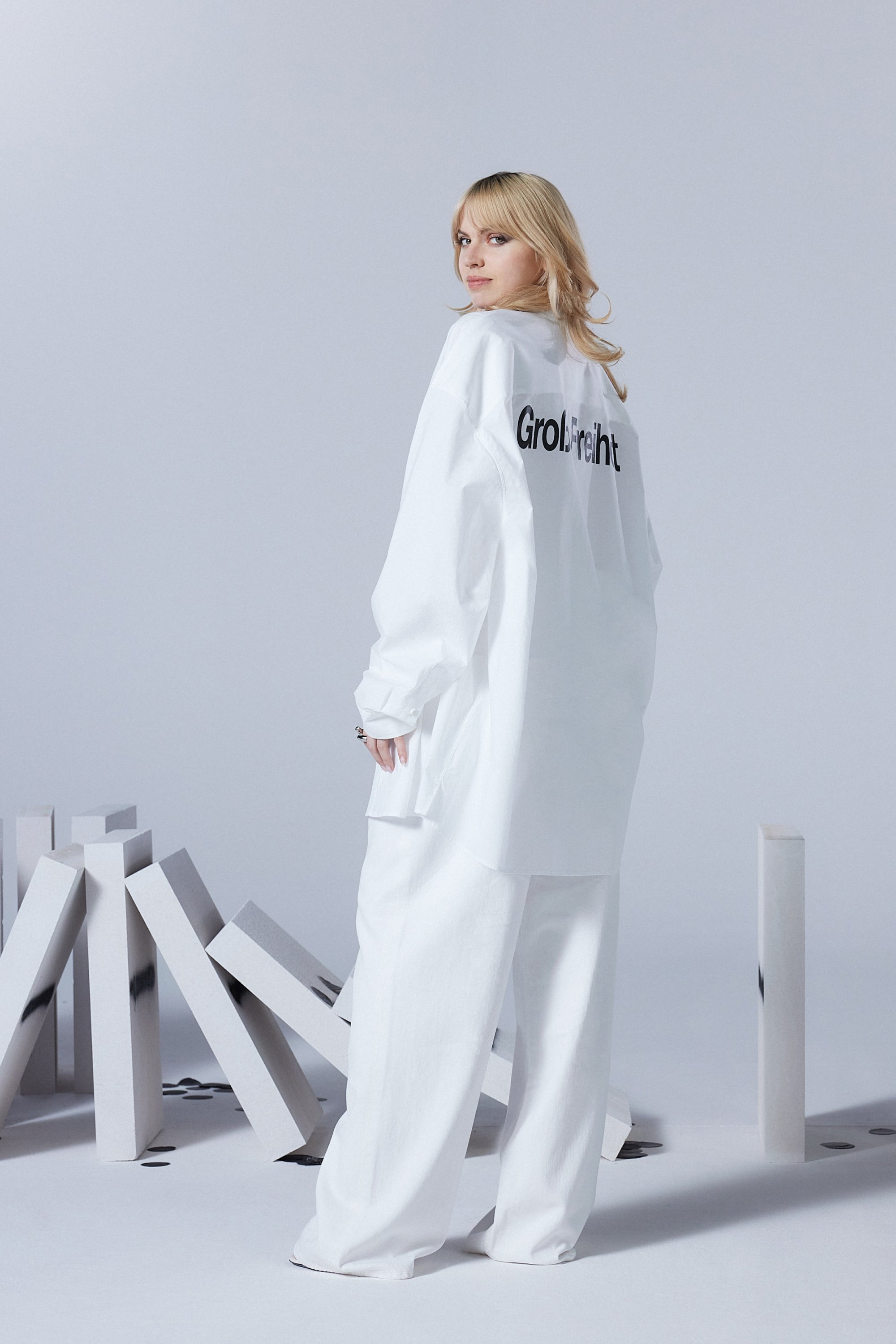A woman with long hair looks back while wearing an oversized white outfit labeled "Grolfreight," standing near scattered white objects.