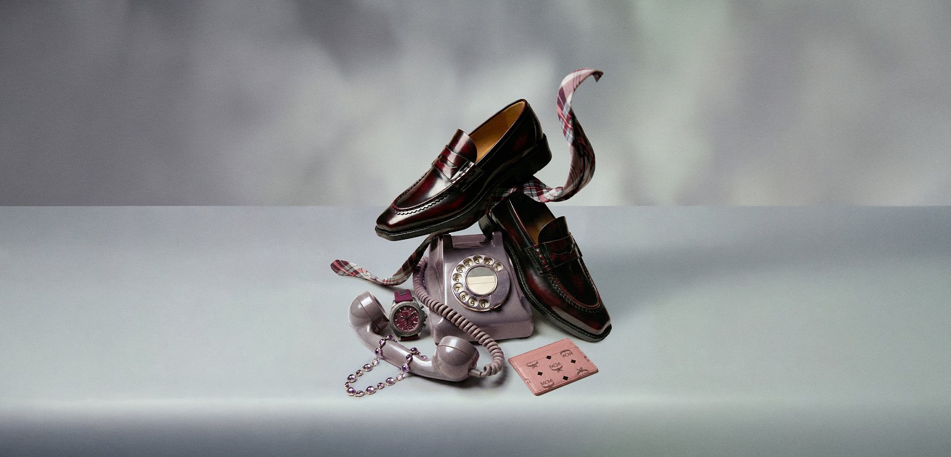 Par de mocasines de cuero burgundy equilibrados sobre un teléfono vintage de disco, acompañados por un reloj de pulsera, una pulsera de cuentas y un tarjetero rosa.