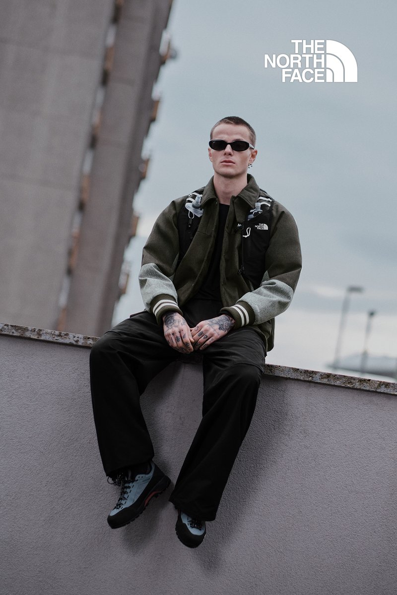 Mørkegrøn og sort jakke med grå ærmer, sorte bukser og blå og sorte sko. Tatoveringer synlige på hænderne, iført solbriller.