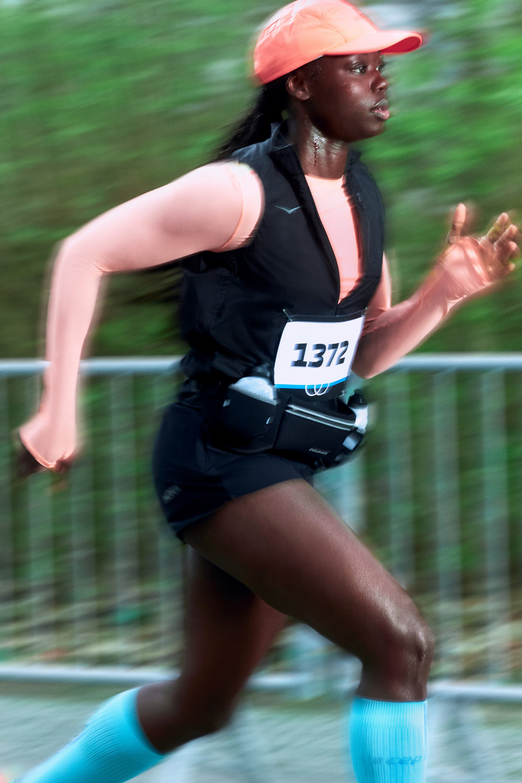 A runner wearing a black vest and a pink cap races vigorously, with a bib number visible, amidst a blurred natural background.