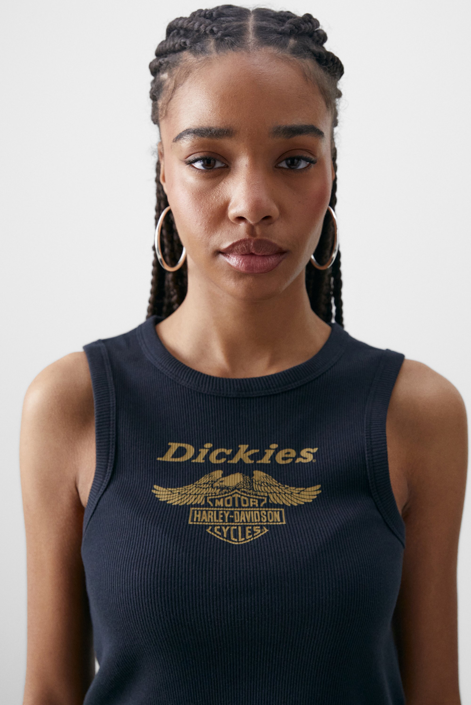 Een jonge vrouw met gevlochten haar draagt een donkere mouwloze top met een gouden grafisch ontwerp dat de woorden "Dickies" en motorrijdersafbeeldingen bevat.
