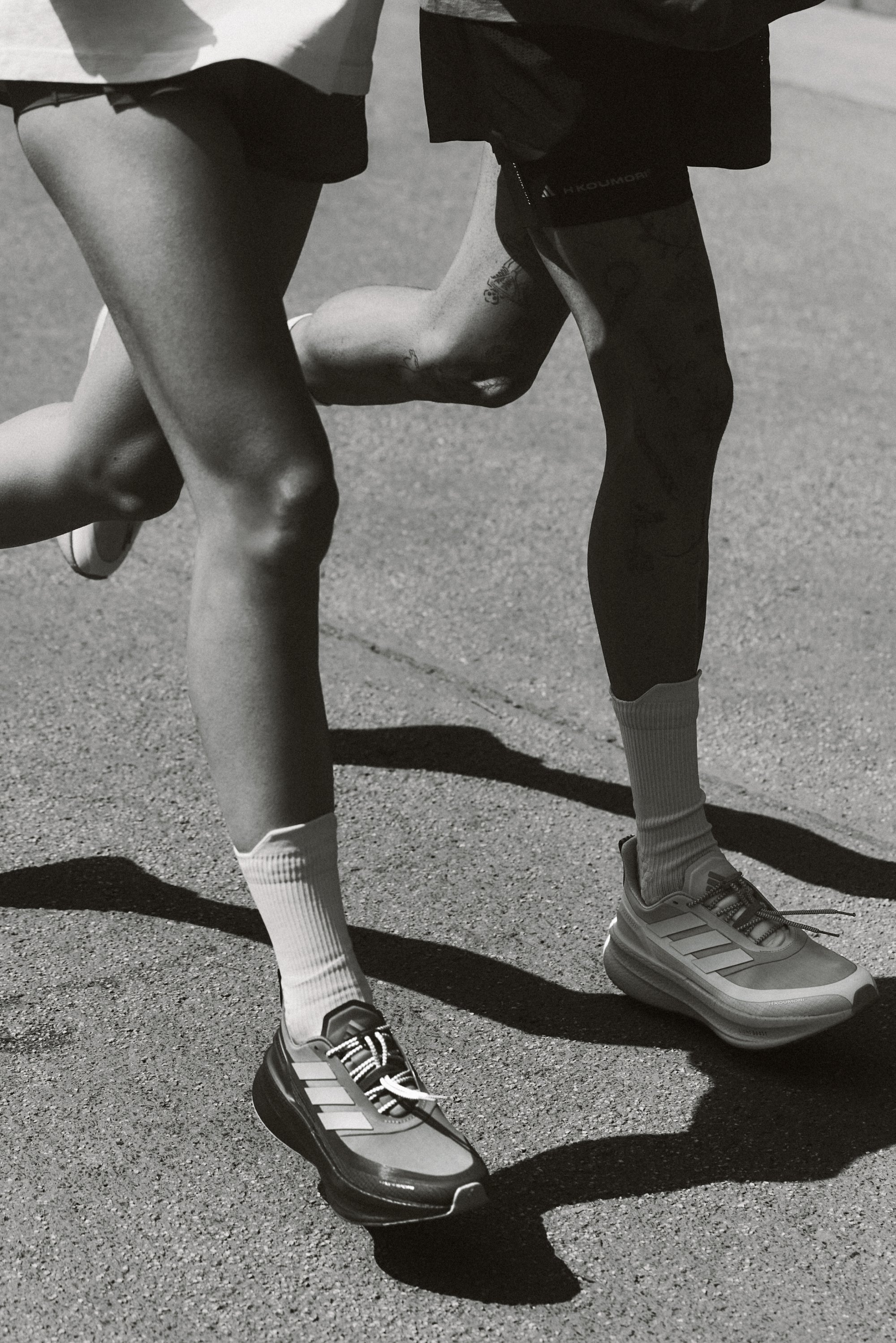 Due paia di gambe che corrono su una strada, indossando scarpe da ginnastica e calze bianche. La scena è in bianco e nero, evidenziando il movimento e le ombre.